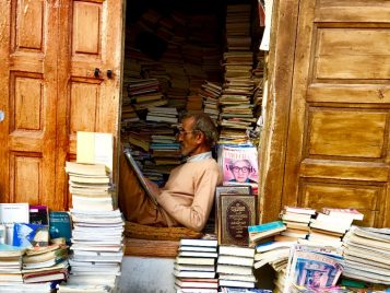 A man sits in a distant doorway that is surrounded by stacks of books inside and out. The man is reading.